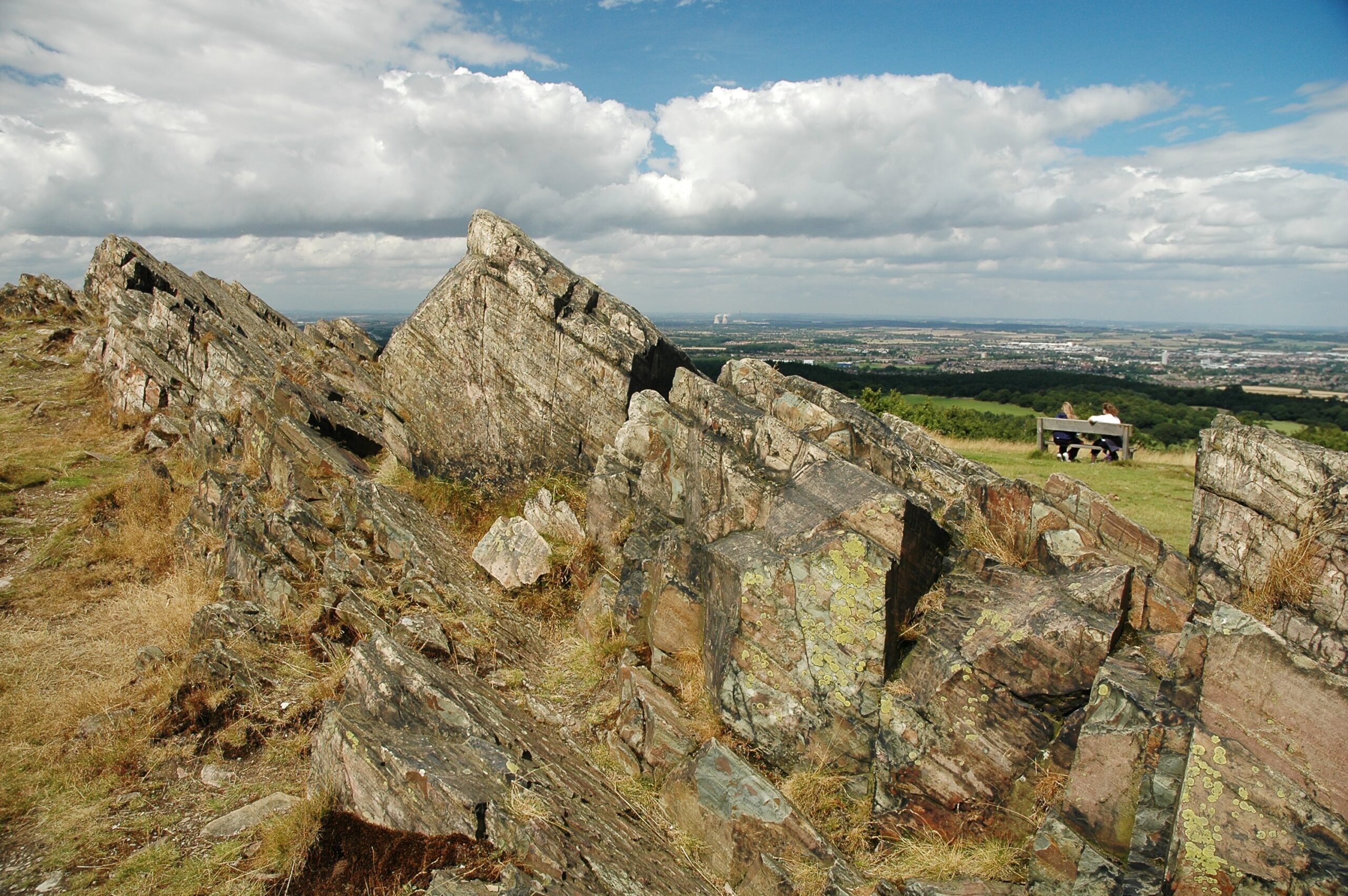 Inclined layers of rock at Beacon Hill. In the middle distance two people sit on a bench admiring the view over Loughborough.