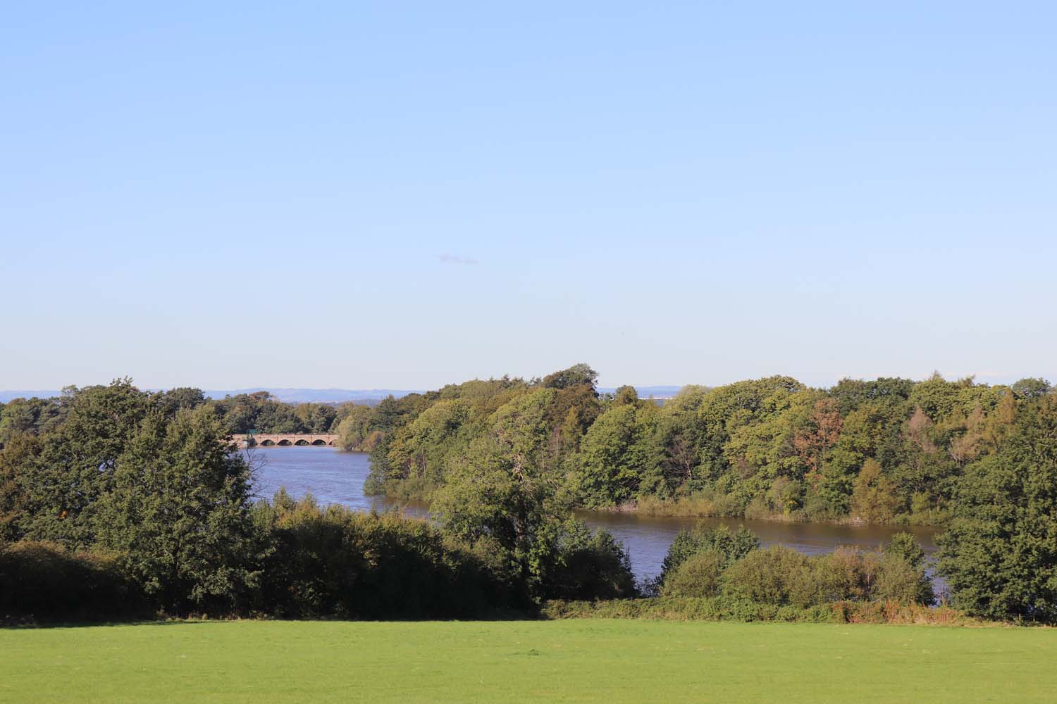 A view over Blackbrook Reservoir. The water is surrounded by trees. A bridge is visible in the distance.