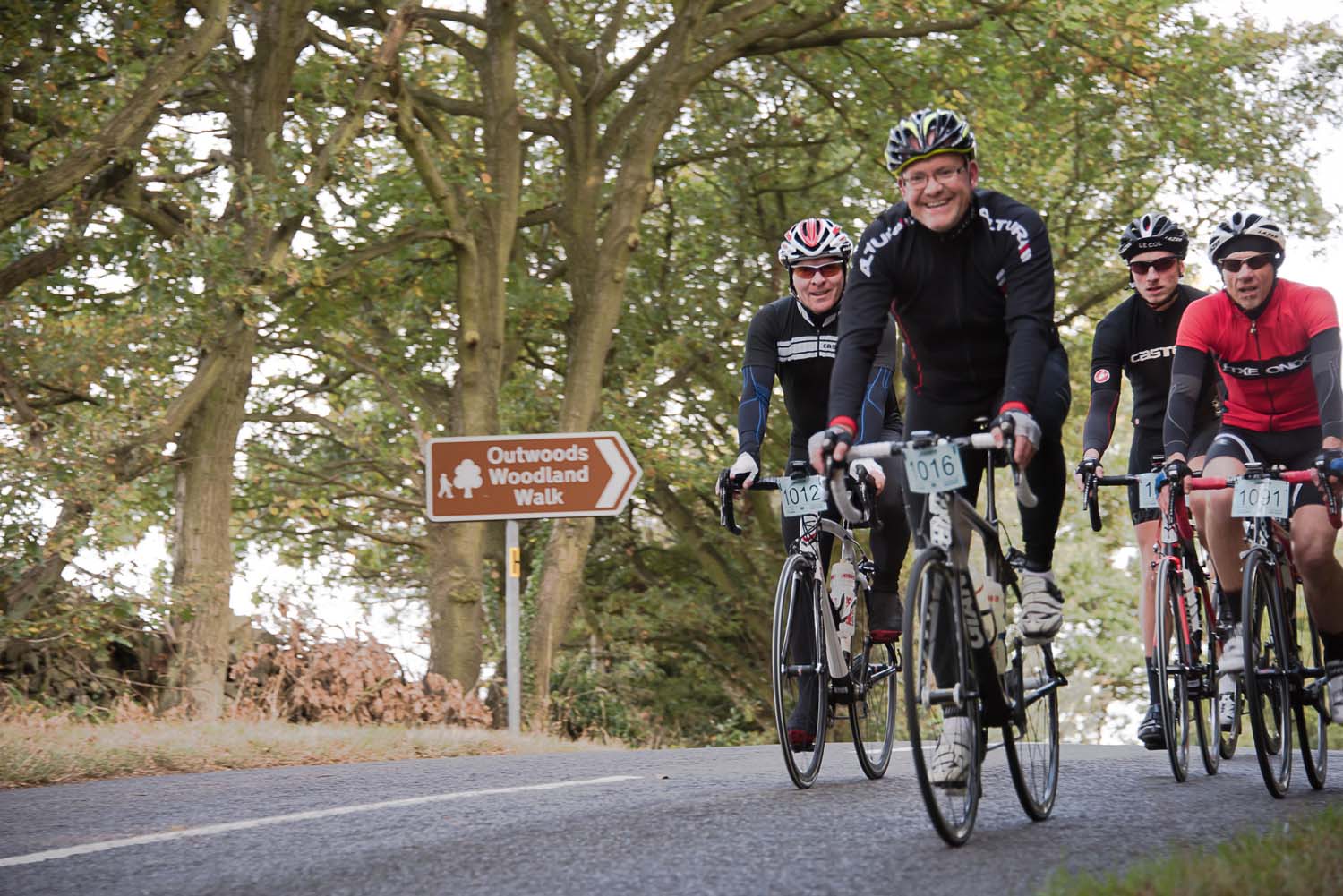 A group of cyclists rides past a sign saying "Outwoods Woodland Walk"