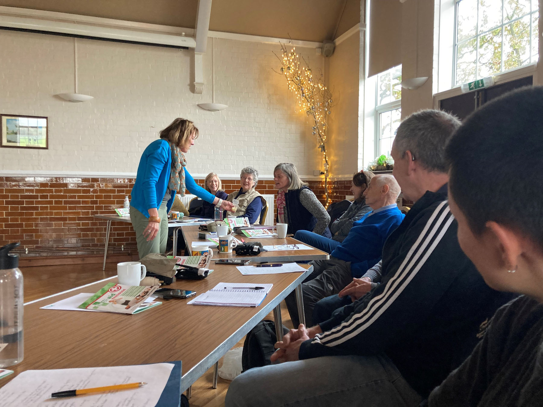 A woman provides first aid training to a group of around 8 people in a village hall