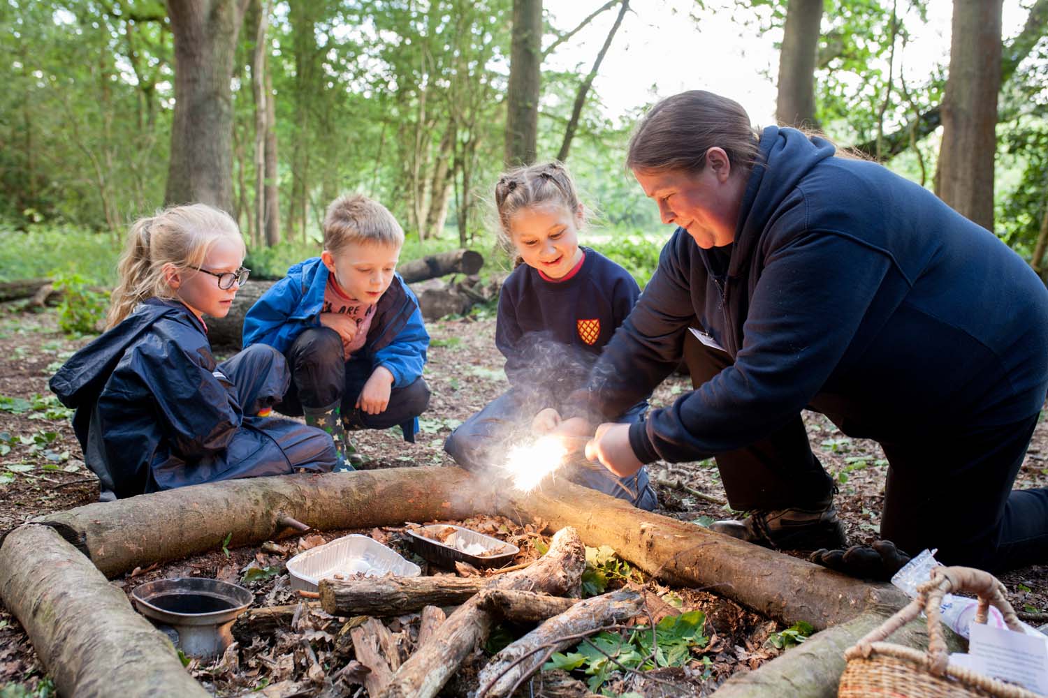 An instructor demonstrates to three children how to start a fire using a fire stick, as part of a Forest School