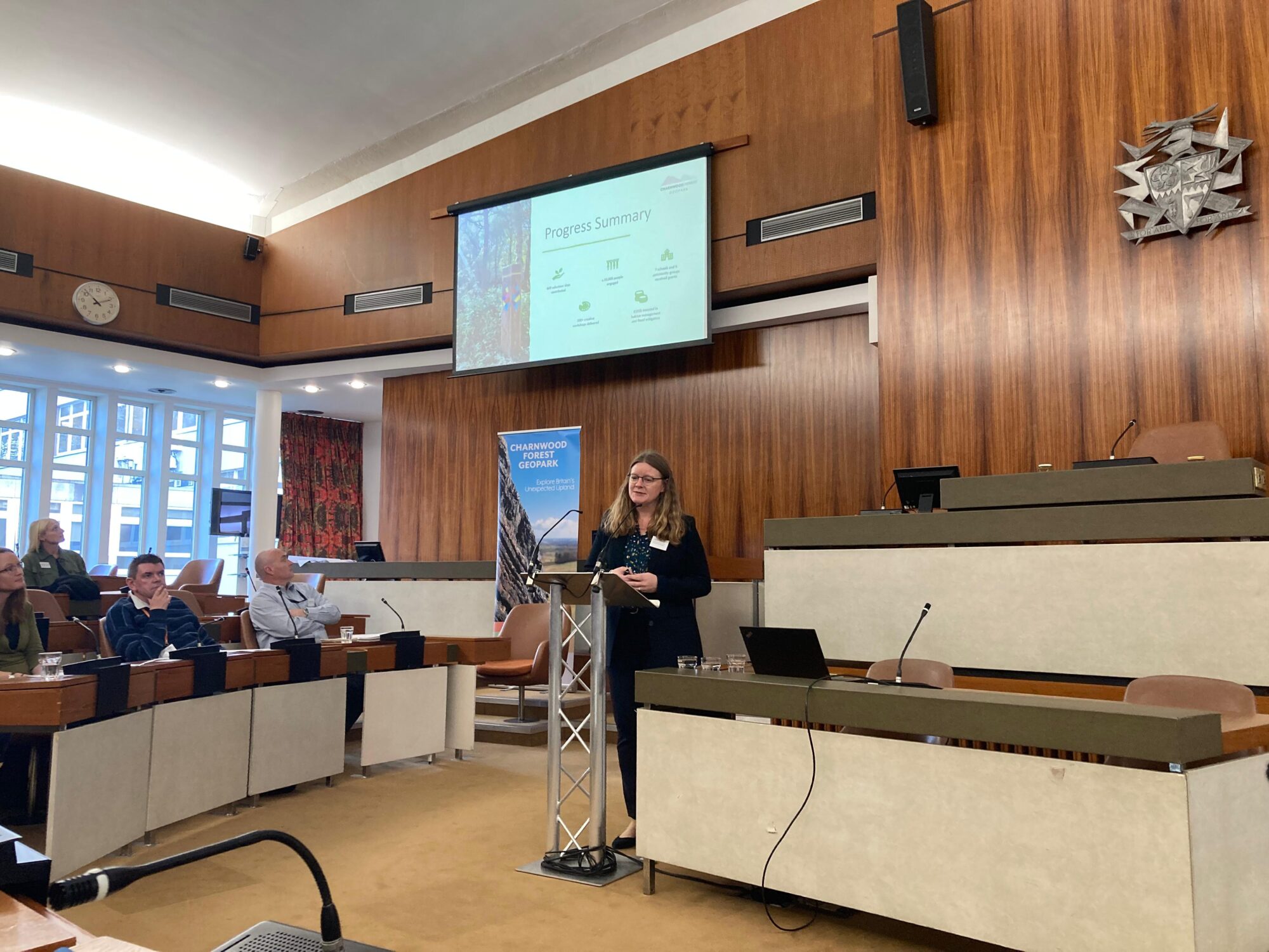 A presenter is speaking in front of a screen that says "Progress Summary"